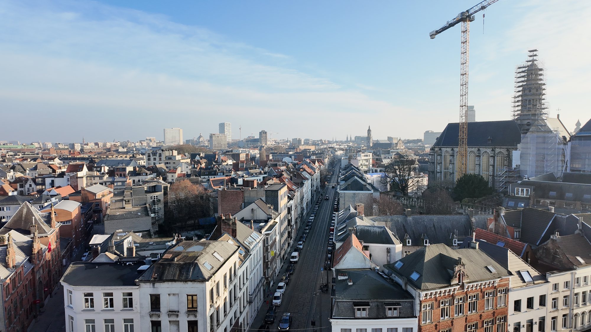 Elevated view over Antwerp's rooftops toward a Gothic church spire wrapped in construction scaffolding, with a tram line running down the street below