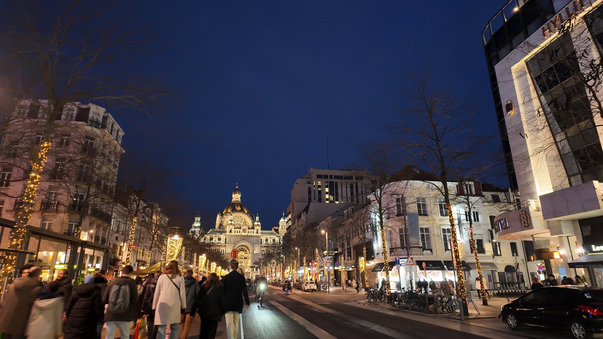 The street leading to Antwerp Centraal Station at dusk, strung with holiday lights, crowds walking toward the station's illuminated dome in the distance