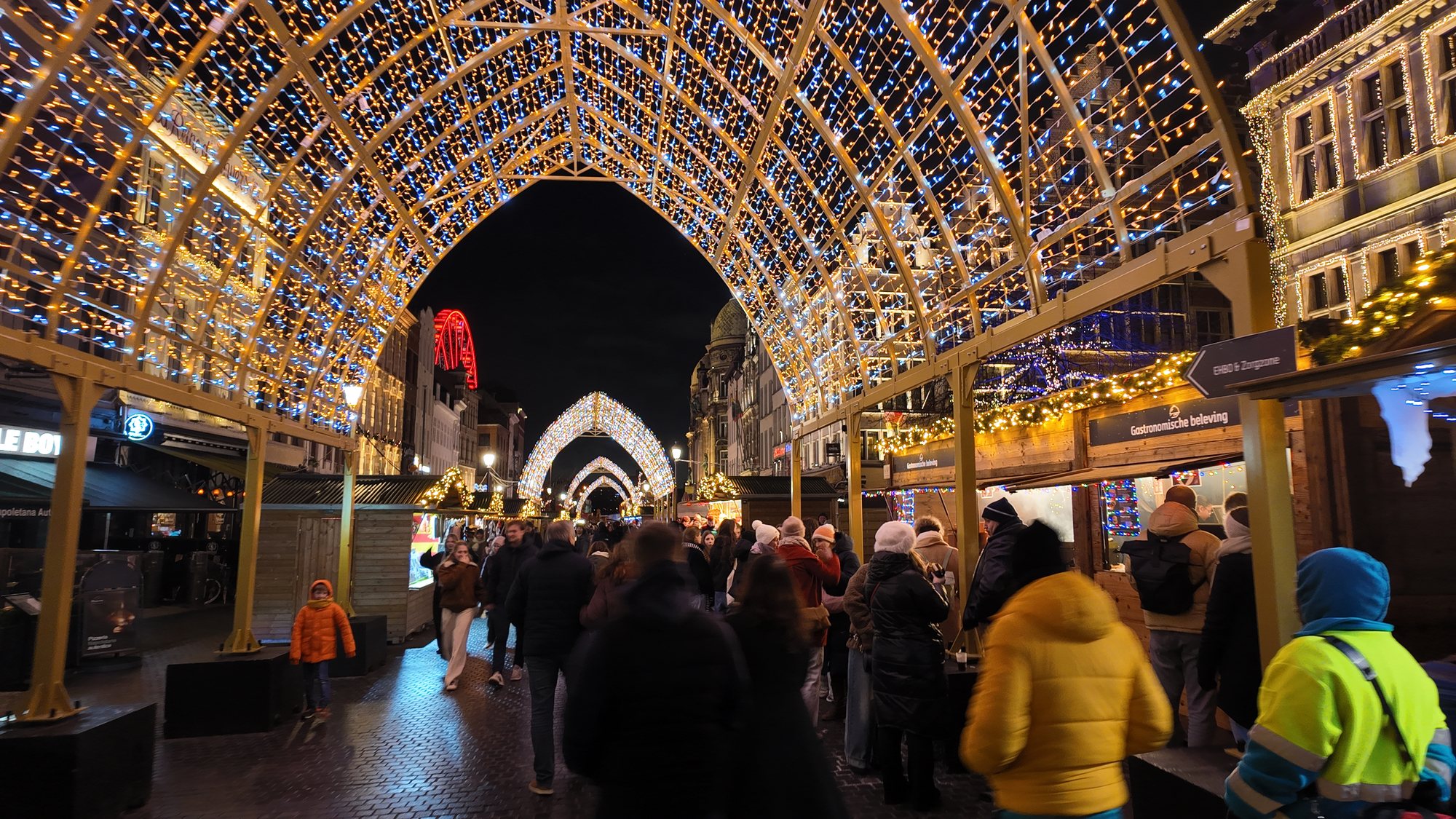 An illuminated arch tunnel over Antwerp's Christmas market stalls, ferris wheel glowing red in the background